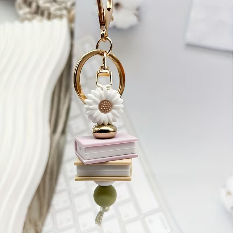 A handcrafted silicone beaded keyring featuring a white sunflower bead, a stack of two miniature book beads (pink and apricot), a gold accent bead, and a natural wood bead on a gold-tone keyring. The keyring is displayed over a white desk and computer keyboard.