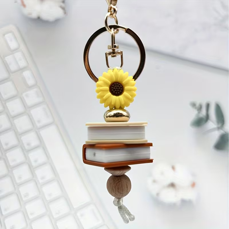 A handcrafted silicone beaded keyring featuring a bright yellow sunflower bead, a stack of two miniature book beads (cream and brown), a gold accent bead, and a natural wood bead on a gold-tone keyring. The keyring is displayed over a white desk and computer keyboard.