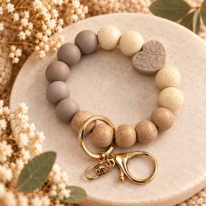 A silicone and wooden bead wristlet keychain from Earthy Heart & Timber Wristlet Keychain.png resting on a round cream stone tray. The beaded loop features smooth taupe silicone beads, natural wood beads, cream-colored silicone beads textured with delicate lace filigree patterns, and a taupe heart-shaped silicone focal bead engraved with a floral botanical motif. A gold key ring and lobster clasp are attached to the wooden bead section