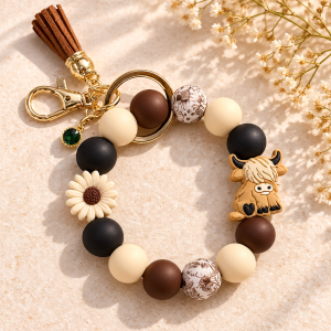 Handmade Highland cow beaded wristlet keychain in mocha brown, black and cream, featuring botanical-print beads, a daisy charm, brown tassel and gold-tone hardware on a warm neutral background.