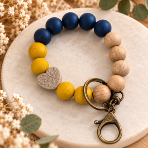 A silicone and wooden bead wristlet keychain from Midnight Navy & Mustard Heart Wristlet.png, displayed on a round cream marble platter. The beaded bracelet features smooth matte navy blue beads, natural light wood beads, textured mustard yellow beads with detailed lace or filigree patterns, and a gray heart-shaped silicone focal bead engraved with a floral botanical design. Attached to a dark antique brass key ring and lobster clasp, the overall look combines bold color contrast with rustic textures.