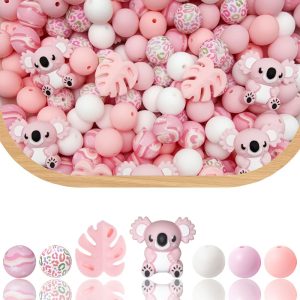 A product display from Pink Koala Leaf 44 Piece.jpg featuring a variety of pastel pink, peach, and white crafting beads. The upper portion shows a wooden bowl overflowing with a mixed pile of loose round and shaped silicone beads. Nestled within the pile are multiple small, pink cartoon koala focal beads and pink monstera leaf focal beads. The lower portion displays a clean, single row of the bead varieties with their reflections on a white surface below them, showcasing a pink marble-swirl bead, a multi-colored leopard print bead, a pink tropical leaf bead, a pink koala character bead, a solid white bead, a solid light pink bead, and a solid soft peach bead.