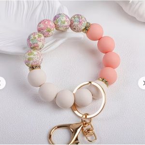 A circular wristlet keychain resting on a white textured surface. The bracelet is made of silicone beads divided into three distinct sections: matte cream beads, soft peach-pink beads, and five patterned beads featuring an intricate pink, green, and purple floral print. The floral section is separated from the solid colors by thin gold spacer rings encrusted with small rhinestones. A polished gold-toned key ring and a matching lobster clasp hang from the bottom of the wristlet.
