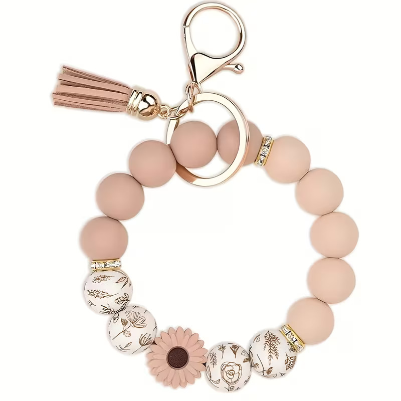 Hand-created beaded wristlet keychain by Woodland Beads & Co featuring matte dusty rose beads, white beads with gold wildflower illustrations, a rose-colored sunflower charm, and a matching tassel with rose-gold hardware.