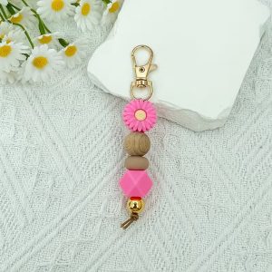 A handmade keychain featuring a pink daisy silicone bead, a wooden bead, a brown silicone bead, a pink geometric bead, and a gold metallic bead, attached to a gold lobster clasp.