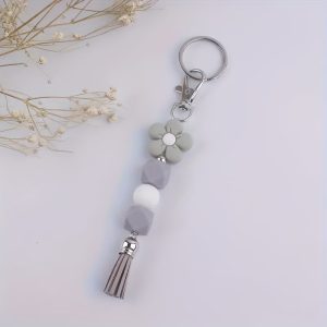 A decorative keychain featuring a slate grey cartoon flower silicone bead, geometric grey beads, a single white bead, and a coordinating grey tassel attached to a silver keyring