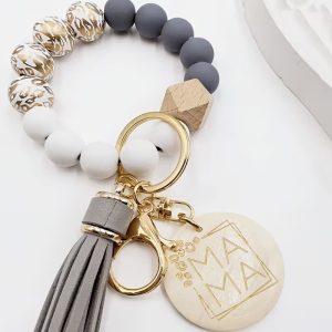 A wristlet keychain from Leopard Print Wristlet.jpg made of silicone and wooden beads arranged in a circle. The beads include matte gray, solid white, a single geometric wooden bead, and white beads with metallic gold leopard print patterns. Attached to a gold key ring and lobster clasp are a long gray faux-leather tassel and a round cream-colored charm engraved with the word "MAMA" inside a thin gold geometric border.