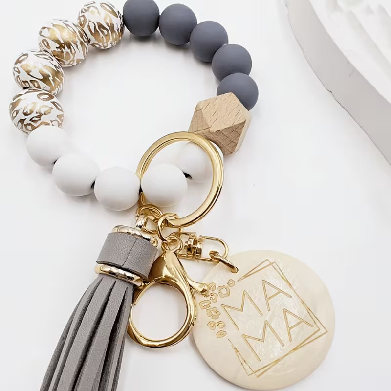 A wristlet keychain from Leopard Print Wristlet.jpg made of silicone and wooden beads arranged in a circle. The beads include matte gray, solid white, a single geometric wooden bead, and white beads with metallic gold leopard print patterns. Attached to a gold key ring and lobster clasp are a long gray faux-leather tassel and a round cream-colored charm engraved with the word "MAMA" inside a thin gold geometric border.