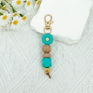 A handmade keychain featuring a teal daisy silicone bead, a wooden bead, a neutral silicone bead, a teal geometric bead, and a gold metallic bead, attached to a gold lobster clasp.