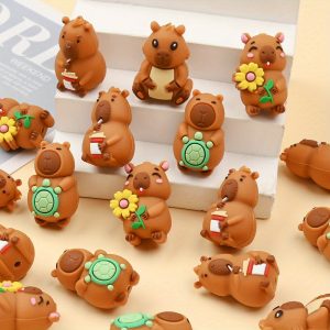 A collection of brown cartoon capybara silicone focal beads scattered across a cream-colored surface and arranged on a small white tiered display stand. The detailed beads feature four distinct capybara designs: one sitting and sipping from a white and red takeaway coffee cup with a straw, one smiling with rosy pink cheeks and holding a yellow sunflower, one standing while holding a small green turtle on its belly, and one sitting forward with a cream-colored belly.