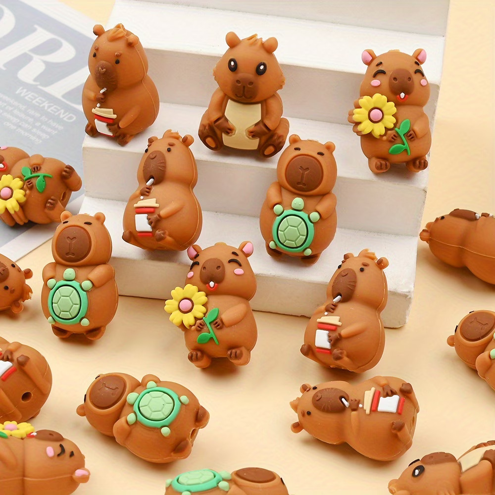 A collection of brown cartoon capybara silicone focal beads scattered across a cream-colored surface and arranged on a small white tiered display stand. The detailed beads feature four distinct capybara designs: one sitting and sipping from a white and red takeaway coffee cup with a straw, one smiling with rosy pink cheeks and holding a yellow sunflower, one standing while holding a small green turtle on its belly, and one sitting forward with a cream-colored belly.