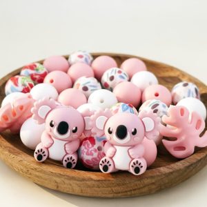 A 34-piece DIY silicone bead kit displayed on a round wooden tray, featuring cartoon pink koala focal beads, pink Monstera leaf shapes, solid pink and white round beads, and patterned beads with red floral and abstract watercolor prints.