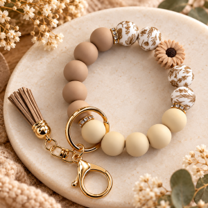 A silicone bead wristlet keychain in warm neutral shades resting on a round cream stone plate. The beaded bracelet features matte taupe and cream round silicone beads, white round beads patterned with gold line-art botanical prints, crystal rhinestone spacer rings, and a small beige silicone sunflower bead. Attached to a gold key ring and lobster clasp is a matching taupe-beige faux-leather tassel with a shiny gold cap.