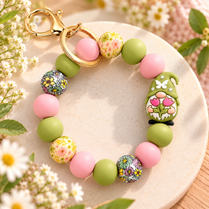 A silicone bead wristlet keychain from Pastel Floral Gnome Wristlet Keychain.png resting on a round stone platter surrounded by white flowers. The wristlet features matte avocado green and light pink round beads, patterned beads printed with yellow, pink, and green flowers, and an adorable silicone garden gnome charm wearing a green hat while holding pink tulips. A shiny gold key ring and lobster clasp are attached at the top.