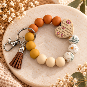 A wristlet keychain made of silicone beads arranged in a circle resting on a round marble tray. The beads include matte terracotta orange, mustard yellow, cream, and white beads patterned with fine green leaves and orange flowers. A beige heart-shaped silicone focal charm is inscribed with the phrase "BE KIND" surrounded by tiny pink flowers. Attached to a silver key ring and lobster clasp is a short brown faux-leather tassel.